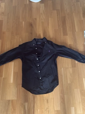 Svart skjorta från Polo Ralph Lauren - Klassisk svart skjorta från Polo Ralph Lauren med lila broderad logga på bröstet. Skjortan har knappar framtill, button-down krage och soprena linjer. Perfekt till jeans eller chinos för en clean look.
