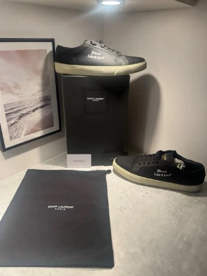 Svarta Saint Laurent sneakers - Snygga svarta sneakers från Saint Laurent med vit logga på sidan och kontrasterande vit sula. Klassisk låg modell med rund tå och snörning. Tillverkade i canvas och skinn för en exklusiv känsla. Perfekta för dig som gillar stilrena och tidlösa sneakers.