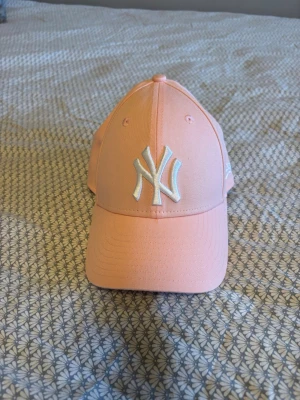 Ljusrosa New Era Yankees keps - Säljer en ljusrosa keps från New Era med New York Yankees-logga broderad i vitt framtill. Klassisk böjd skärm och ventilerande hål upptill. Kepsen är tillverkad i ett slitstarkt tyg och har New Era-loggan broderad på sidan. Perfekt för dig som vill ha en clean och sportig look.