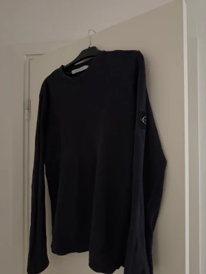 Svart långärmad tröja Calvin Klein - Snygg svart långärmad tröja från Calvin Klein Jeans med diskret struktur i tyget och en stor logotyp-patch på ena ärmen. Tröjan har rund halsringning och är perfekt för en stilren och avslappnad look.