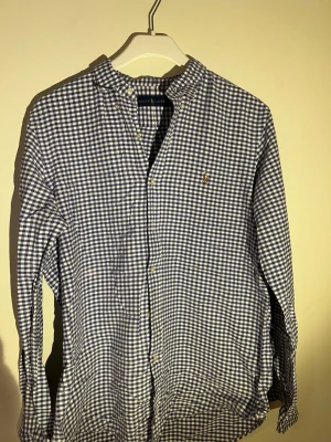 Rutig blåvit skjorta från Ralph Lauren -  beingEn klassisk rutig skjorta från Ralph Lauren i blått och vitt. Skjortan har lång ärm, knappar framtill och en liten broderad logga på bröstet. Tillverkad i mjuk bomull och perfekt för en avslappnad men ändå stilren look.