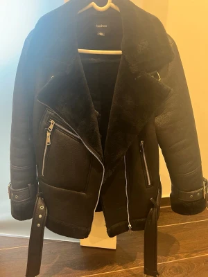 Svart biker-kappa från Boohoo - Snygg svart biker-inspirerad kappa från Boohoo med fuskpälsfoder, bred krage och detaljer i silverfärgad metall. Jackan har dragkedjor, bälten vid ärmslut och nederkant samt två snedställda fickor framtill. Perfekt för dig som vill ha en edgy och varm look.