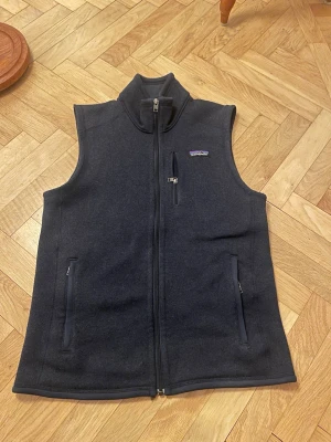 Mörblå fleeceväst från Patagonia - Mörkblå fleeceväst från Patagonia med krage och hel dragkedja framtill. Västen har tre fickor med dragkedja och en liten logga på bröstet. Perfekt till våren 