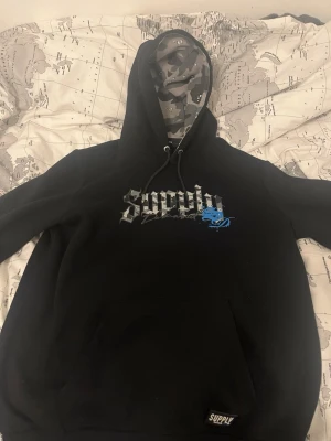 Svart hoodie från Supply med camo-huva - Svart hoodie från Supply med stor magficka och tryck i vitt och blått på bröstet. Huvan har ett grått camomönster på insidan och justerbara snören. Perfekt för en streetwear-look och riktigt skön att bära.