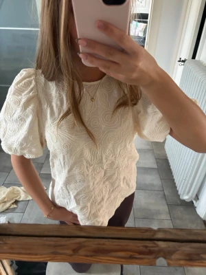 Vit broderad blus med puffärm - Säljer en vit blus med puffiga korta ärmar och vackert broderat mönster över hela tyget. Blusen har knäppning bak och är gjord i ett lätt, strukturerat material som ger en lyxig känsla. Perfekt för dig som gillar unika detaljer och romantisk stil.