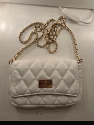 Vit liten handväska med axelband  - Vit lite  väska med gulddetaljer och axelband, från Glitter. Man får lagom plats med telefonen, nycklar och läppglanset🥰Finns ett litet sidofack inuti väskan. Använd några gånger och fortfarande i väldigt fint skick!