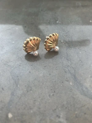 Guldpläterade snäckformade örhängen - Säljer ett par örhängen i snäckform med guldpläterad yta och en liten vit pärla längst ner. De har en stilren och trendig design som ger en lyxig känsla. Perfekta för dig som gillar havsinspirerade accessoarer och vill ha något unikt.