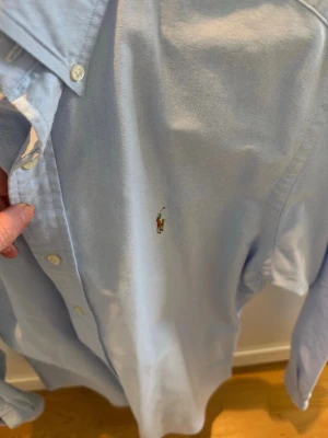 Ljusblå skjorta från Polo Ralph Lauren - Klassisk ljusblå skjorta från Polo Ralph Lauren med broderad logga på bröstet. Skjortan har knappar framtill, button-down krage och långa ärmar. Tillverkad i mjuk bomull för en bekväm känsla.