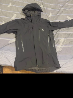 Svart regnjacka från Haglöfs - Snygg svart regnjacka från Haglöfs med justerbar huva och flera vattentäta dragkedjefickor framtill. Jackan har hög krage, långa ärmar och är tillverkad i ett slitstarkt, vattenavvisande material. Perfekt för blöta dagar och outdoor-äventyr. Helt ny utan tags använts en gång