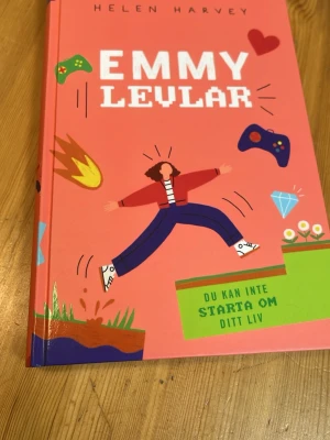 Emmy Levlar av Helen Harvey - En färgstark bok med rosa hård pärm, illustrationer av tv-spel, eldklot och diamant. Boken handlar om Emmy som älskar datorspel och möter utmaningar både i spelet och i verkliga livet. Perfekt för unga som gillar gaming och äventyr.