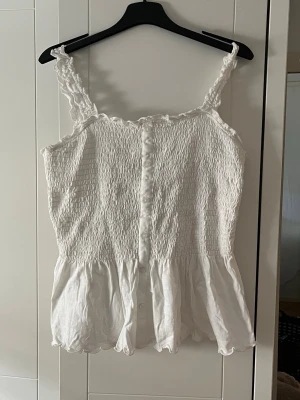 Vit smockad linnetopp med knappar - Söt vit linnetopp med smockad överdel och volangkant nedtill. Toppen har breda axelband med volangdetaljer och knappar framtill. Perfekt för en somrig look och lätt att matcha med jeans eller kjol.