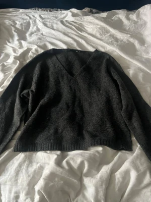 Mörkgrå stickad V-ringad tröja - Mysig mörkgrå stickad tröja med V-ringning och långa ärmar. Tröjan har en rak passform och ribbstickade muddar vid ärmslut och nederkant. Perfekt för lager-på-lager under kyliga dagar.