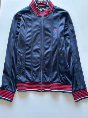 Ben Sherman Track Jacket – Vintage British Casual Style – S - Marinblå track jacket från Diadora med röda och vita ränder på krage, ärmslut och midja. Jackan har dragkedja framtill, två fickor med dragkedja och broderad Diadora-logga på bröstet. Tillverkad i glansigt syntetmaterial för en sportig retrostil. 📏 Mått Axel till axel  – 61 cm Axel till nederkant – 70 cm