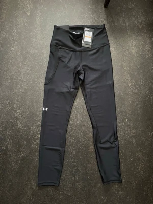 Svarta leggings/ tränings byxor från Under Armour - Snygga svarta leggings från Under Armour med hög midja och praktiska sidofickor. Materialet är stretchigt och passar perfekt för träning eller chill. Diskret logga på benet och modern passform som sitter tight. Perfekt för dig som gillar sportiga och bekväma kläder.