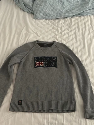Grå stickad tröja Morris - Snygg grå stickad tröja från Morris med broderad text 'Morris 1968' och brittiska flaggan på bröstet. Tröjan har rund hals, långa ärmar och ribbade muddar vid ärmslut och nederkant. Perfekt för dig som gillar klassisk och stilren design.