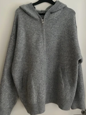 Grå hoodie med dragkedja - Mysig grå hoodie med huva och halv dragkedja framtill. Tillverkad i mjukt stickat material och har två fickor på sidorna. Perfekt för en avslappnad och chill stil.