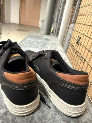 Svarta loafers från Lovve - Säljer ett par svarta loafers från Lovve med vita sulor och bruna detaljer vid hälen. Skorna har en klassisk låg profil och är tillverkade i ett slitstarkt material med snygga sömmar. Perfekta för en stilren och avslappnad look.