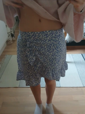 Blommig omlottkjol med volang - Supersöt kort kjol med blått blommönster på vit botten. Kjolen har omlottmodell med knyt i sidan och volang längs nederkanten. Perfekt för dig som gillar en feminin och trendig stil.