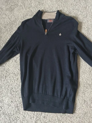 Marinblå ziptröja från Morris - Snygg marinblå tröja från Morris med halv dragkedja och ribbade muddar. Tröjan har en liten broderad logga på bröstet och kontrastfärgad insida vid kragen. Perfekt till jeans eller chinos för en stilren look.