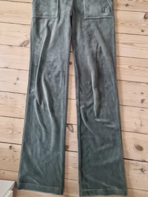 Grå velourbyxor från Juicy Couture - Mjukisbyxor i grön velour från Juicy Couture med raka ben och bred resår i midjan. Byxorna har snörning och stora fickor framtill, samt en broderad JC-logga på ena fickan. 