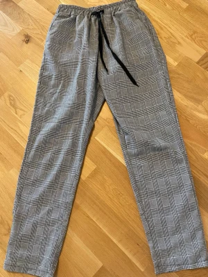 Rutiga mjukisbyxor med dragsko - Säljer ett par gråa mjukisbyxor med svartvitt rutmönster och svart dragsko i midjan. Byxorna har avslappnad passform och resår i midjan för extra komfort. Perfekta för chill dagar eller när du vill vara bekväm men ändå se stilren ut. Använd en enda gång 