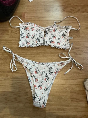 Blommig bikini med volang och knyt - Supersöt bikini i vitt med blommigt mönster i rosa.. Överdelen har volangkant, smala axelband och knyt bak. Underdelen har högt skurna ben och knyt i sidorna. Perfekt för sommaren!