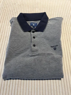 Grå långärmad skjorta från Gant - Storlek:M, normal i storleken.                                Grå långärmad tröja från Gant med mörkblå krage och tre knappar framtill. Tröjan har diskret mönster och Gant-logga broderad på bröstet. Perfekt för dig som gillar stilrena och klassiska plagg.