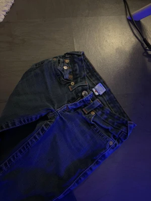 True Religion blå jeans med fickdetaljer - Säljer ett par blå jeans från True Religion med ikoniska kontrastsömmar och snygga ficklock bak. Jeansen har raka ben och klassisk femficksdesign. Materialet är slitstark denim i bomull. Perfekta för dig som gillar streetwear och vill sticka ut med detaljer.