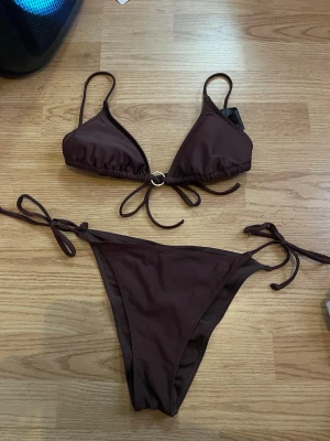 Brun bikini med knyt och ringdetalj - Snygg brun bikini med trekantsöverdel och knyt både vid rygg och höfter. Överdelen har en metallring i mitten för extra detalj. Klassisk modell som passar perfekt till stranden eller poolen.