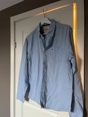 Ljusblå skjorta från Hollister - Säljer en klassisk ljusblå skjorta från Hollister med button-down krage och bröstficka med liten logga. Skjortan har långa ärmar och stängs med knappar framtill. Perfekt till jeans eller chinos för en clean och enkel stil.