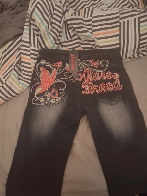 Mörkblå jeans med brodyr från Romwe - Unika mörkblå jeans från Romwe med färgglad brodyr på bakfickorna, inklusive en stor rosa fjäril och texten 'Rave Breed'. Framtill finns rosa detaljer och en liten broderad blomma. Jeansen har en tvättad look och boot cut. Perfekta för dig som vill sticka ut.
