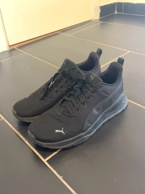 Svarta Puma sneakers mesh - Snygga helsvarta sneakers från Puma med mesh-ovandel och klassisk logga på sidan och hälen. Skorna har lågt skaft, svarta snören och en bekväm sula som passar perfekt till streetwear eller träning. Stilren design som funkar till det mesta.