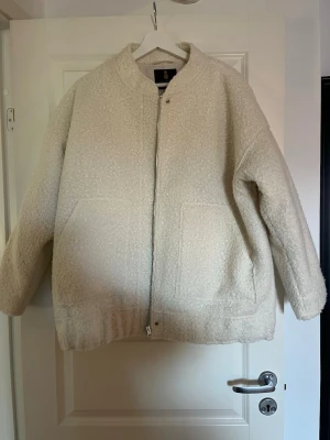 Beige oversized teddyjacka - Mysig oversized teddyjacka i beige med dragkedja framtill och två stora fickor. Jackan har en enkel, kraglös design och elastisk nederkant för en avslappnad look. Perfekt för kalla dagar när du vill vara både varm och trendig.