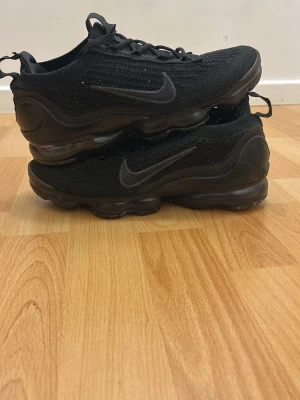Svarta Nike Air VaporMax sneakers - Säljer ett par helsvarta Nike Air VaporMax sneakers med stickad ovandel och synlig Air-sula. Skorna har en låg profil, klassisk Nike Swoosh på sidan och snörning framtill. Perfekta för dig som gillar sportig och stilren look.