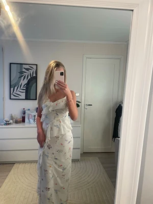 Vit långklänning med blommigt mönster - Säljer en vit långklänning med blommigt mönster och volangdetaljer. Klänningen har v-ringning och är ärmlös med smala axelband. Perfekt för sommarens tillfällen och har en romantisk känsla. Materialet känns lätt och luftigt. Endast använd 1 gång.💕