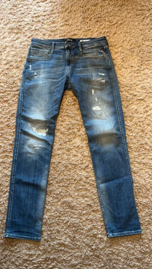 Blå jeans från Replay - Säljer ett par blå jeans från Replay med slitningar och distressed detaljer på framsidan. Modell anbass storlek 34 