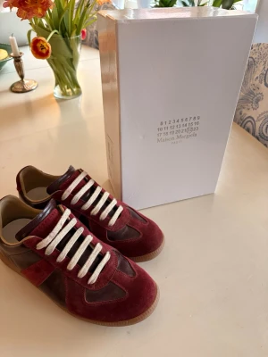 Maison Margiela vinröda gats - 🌟Snygga och stilrena sneakers från Maison Margiela i vinröd mocka och läder. Väldigt clean modell som funkar till det mesta och ger en exklusiv look.  🌟I fint skick och säljs med originalkartong, dustbags samt extra nya skosnören.  🌟Perfekta om du vill ha något unikt men fortfarande stilrent. Skickas snabbt vid köp!😁