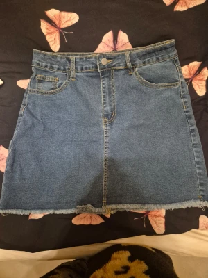 Blå jeanskjol - Säljer en klassisk blå jeanskjol med rå, fransig kant nertill. Kjolen har fem fickor, bälteshällor och stängs med knapp och dragkedja framtill. Perfekt för dig som gillar en avslappnad och trendig stil.