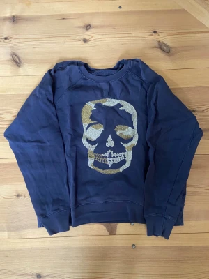 Mörkblå sweatshirt conceptual skull - Cool mörkblå sweatshirt från Zadig & Voltaire med ett stort glittrigt dö,skalle-tryck i guld och silver på bröstet. Tröjan har rund halsringning och ribbade muddar vid ärmslut.