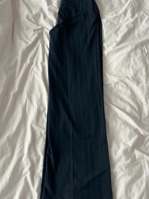 Mörkblå randiga konstymbyxor  - Mörkblå kostymbyxor med diskreta vita kritstrecksränder och raka ben. Byxorna har mellanhög midja, knapp och dragkedja framtill samt sidofickor. Stilrena byxor! Vet tyvärr inte storleken men det borde vara w26 L32/34.