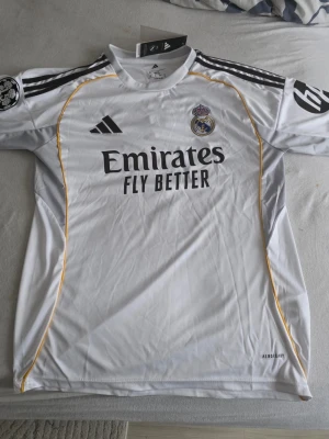 Real Madrid matchtröja Vini Jr #7 - Säljer en vit Real Madrid matchtröja från Adidas med Vini Jr och nummer 7 på ryggen. Tröjan har svarta detaljer camparmar, klubbmärke och sponsortryck framtill samt gula linjer längs sidorna. Tillverkad i halfway precipitation Aeroready-material.