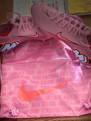 Rosa Nike Mercurial fotbollsskor elite  - Säljer ett par rosa Nike Mercurial fotbollsskor med svart och orange swoosh, samt stora vita och svarta detaljer på hälen. Skorna har snörning och ovandel i textil för bra passform och känsla på planen. Perfekta för dig som vill sticka ut på fotbollsplanen.