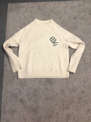 Vit stickad tröja med broderi - Säljer en krämvit stickad tröja med rund hals och ribbade muddar. På bröstet finns ett broderat svart monogram med bokstäverna 'OX'. Tröjan har en avslappnad passform och är perfekt för lager på lager. Materialet känns mjukt och bekvämt.