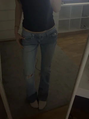 Ljusblå slitna bootcut jeans - Säljer ett par ljusblå bootcut jeans med slitningar på knät och snygga detaljer på bakfickan. Jeansen har låg midja och klassisk femficksdesign. Perfekta för en avslappnad och trendig look. storlek 170 men passar mig som brukar ha s