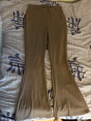 Bruna utsvängda kostymbyxor NLY M - Säljer ett par ljusbruna kostymbyxor från NLY med utsvängda ben och hög midja. Byxorna har bälteshällor och är gjorda i ett mjukt, stretchigt tyg som sitter snyggt. Perfekta för dig som gillar en trendig och bekväm look.