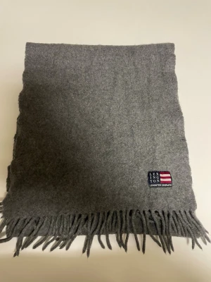 Grå halsduk från Lexington - Klassisk grå halsduk från Lexington med mjukt material och stilren design. Halsduken har fransar i kanterna och en liten Lexington-logga med amerikansk flagga broderad i hörnet. Perfekt accessoar för att hålla värmen och addera stil till din outfit.