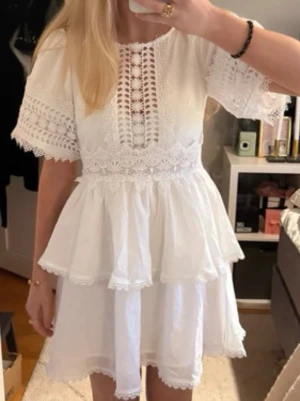 Vit kort klänning med spetsdetaljer - Söt vit kortklänning med volanger och vackra spetsdetaljer på ärmar och överdel. Klänningen har trekvartsärm och omlottliknande spetsmönster framtill. Perfekt för sommarens alla tillfällen och student.