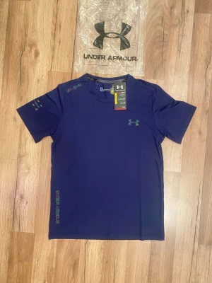 Blå träningströja från Under Armour - Snygg blå t-shirt från Under Armour med rund hals och korta ärmar. Tröjan har diskreta loggor på bröstet, ärmen och sidan samt textdetaljer. Tillverkad i ett lätt och snabbtorkande funktionsmaterial, perfekt för träning.