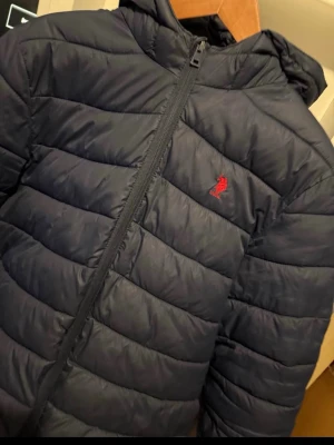 Svart pufferjacka från U.S. Polo Assn. - Svart pufferjacka med huva från U.S. Polo Assn. Jackan har en quiltad look, dragkedja framtill och röd broderad logga på bröstet. Tillverkad i syntetmaterial som håller värmen och ger en clean stil.