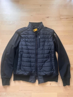 Svart pufferjacka från Parajumpers - Snygg svart pufferjacka från Parajumpers med quiltad front och rygg, mjuka ärmar och hög krage med gul spänndetalj. Jackan har flera dragkedjefickor och en patch på ärmen. Insidan är ljusgrå och jackan har en modern, sportig look.